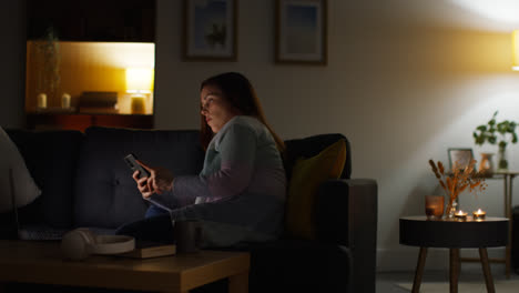 Mujer-Sentada-En-Un-Sofá-En-Casa-Por-La-Noche-Transmitiendo-O-Viendo-Una-Película-O-Programa-En-Una-Computadora-Portátil-Y-Navegando-Por-Internet-En-El-Teléfono-14